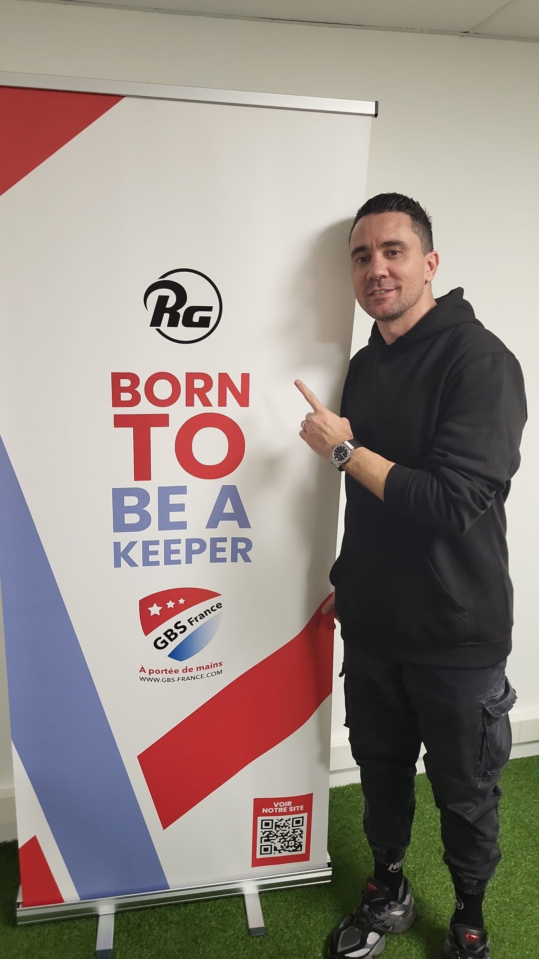 Ramiro Gonzales en tenue décontractée pointe une affiche RG marquée "Born to be a Keeper", représentant une marque d’équipement de gardiens